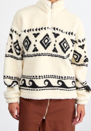 Fleece trui met zwarte geometrische en spiraalvormige patronen op een crèmekleurige achtergrond, halve rits aan de voorkant, gedragen met een bruine rok met ritssluiting voor.