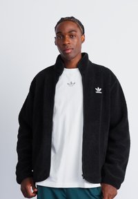 Zwarte fleece jas met een ritssluiting aan de voorkant, hoge kraag en geborduurd logo op de borst. Draagt over een wit shirt voor contrast.