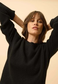 Czarny sweter z dzianiny o okrągłym dekolcie i luźnych rękawach. Miękka tekstura z prążkowanymi mankietami. Modelka pozuje z rękami za głową.
