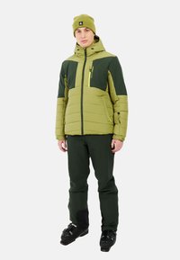 Zelena i tamnozelena izolirana skijaška jakna s kapuljačom, zipom na džepovima i elastičnim manžetama, uparena s odgovarajućim skijaškim hlačama i crnim čizmama.