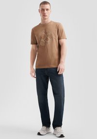 Camiseta marrón de manga corta con un gráfico de logotipo circular. Llevada con jeans azul oscuro y zapatillas blancas sobre un fondo neutro.