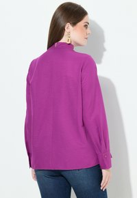 Blusa a maniche lunghe di un vivace viola, con uno spacco sulla schiena, scollo rotondo e polsini con bottoni. Tessuto morbido e vestibilità comoda.
