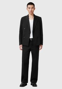 Giacca da suit nera su misura, doppiopetto, con colletto elegante e tasche frontali, abbinata a pantaloni larghi coordinati e scarpe nere.