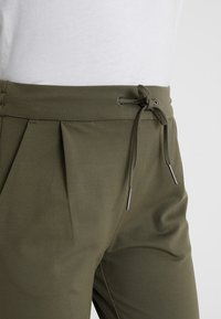 Olivegrüne Hose mit Kordelzugbund, ausgestattet mit Falten und silbernen Metallakzenten an den Kordeln. Hergestellt aus strukturiertem Stoff.