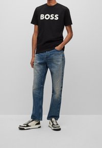 Schwarzes Baumwoll-T-Shirt mit "BOSS" in Weiß bedruckt. Hellblaue, abgenutzte Jeans. Schwarz-weiße Sneaker mit kontrastierenden Akzenten.