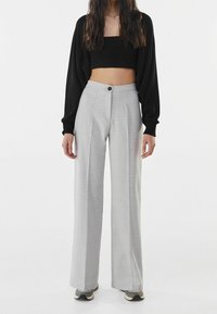 Kobieta w szerokich, szarych, w kratkę spodniach o wysokim stanie, czarnej, krótkiej bluzce z długim rękawem i szarych trampkach stoi na tle białym.