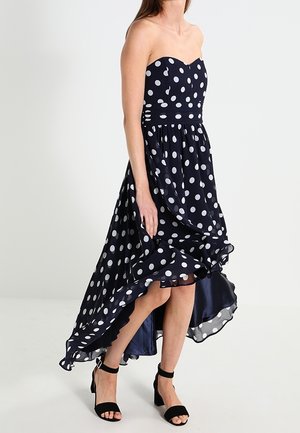 Strapless navy blue dress with white polka dots, high-low hem, worn with black ankle-strap block heel sandals, model walking.