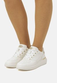 Weiße Sneakers aus strukturiertem synthetischem Material, Schnürdesign, goldene Akzente an den Ösen und der Ferse sowie eine dicke weiße Sohle.