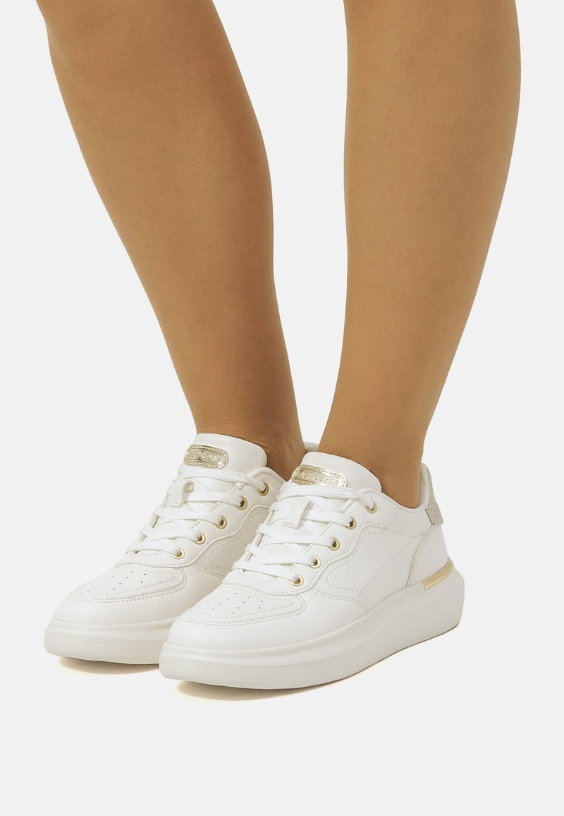 Weiße Sneakers aus strukturiertem synthetischem Material, Schnürdesign, goldene Akzente an den Ösen und der Ferse sowie eine dicke weiße Sohle.
