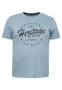 Gaiši zila kokvilnas T-kreklu ar īsām piedurknēm, apaļu kaklu un melnu drukātu tekstu, kas norāda "Heritage Blend" un "Blue Denim" uz priekšpuses.