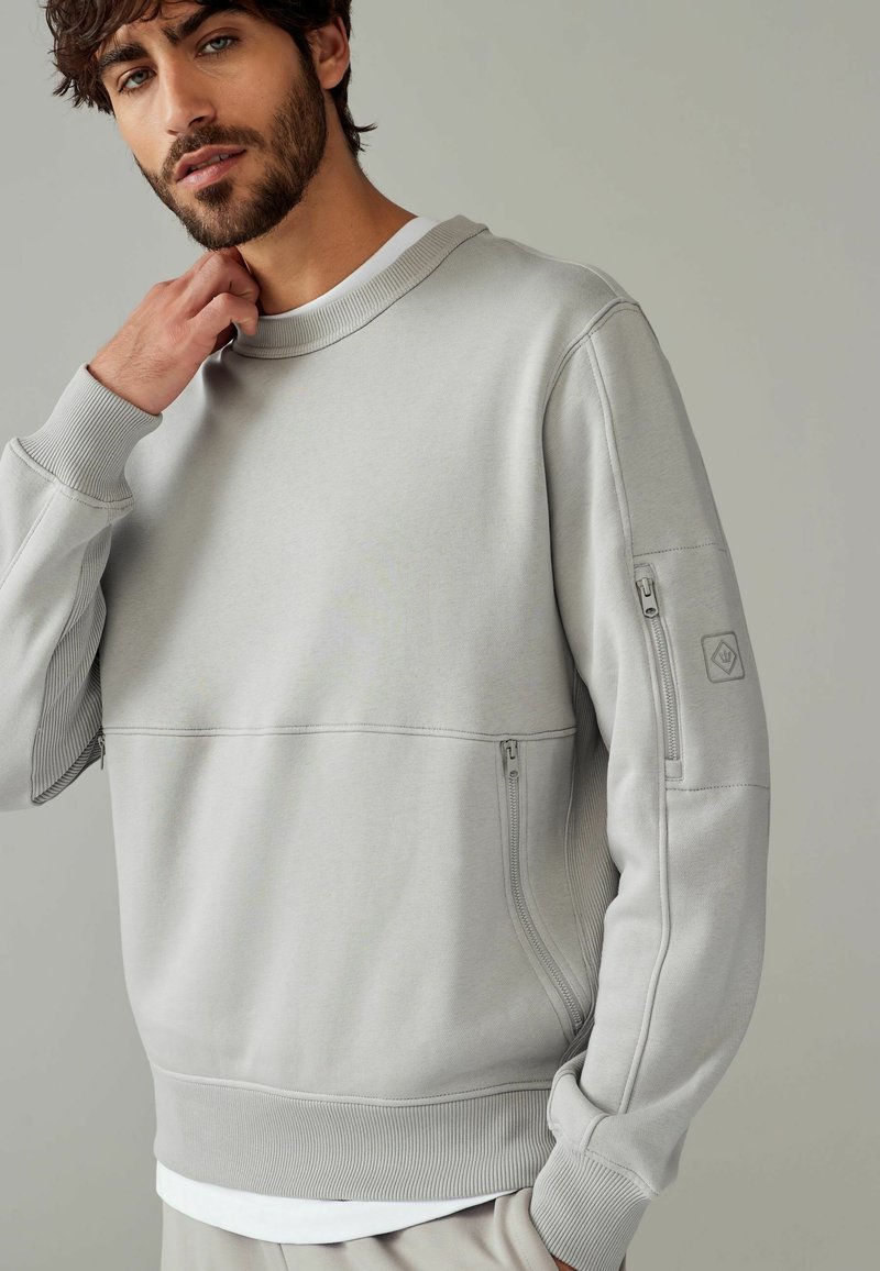 Lys grå sweatshirt med rund halsudskæring og lynlåslomme i siden, lavet af blødt stof med ribbede manchetter og kant.