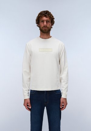 Uomo con capelli ricci e barba che indossa una camicia bianca a maniche lunghe con un piccolo logo e jeans blu, in piedi contro uno sfondo semplice.