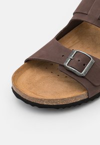Sandal in camoscio marrone con una larga fascia e fibbia in metallo argentato, soletta in sughero, superficie testurizzata di colore marrone chiaro e suola in gomma nera.