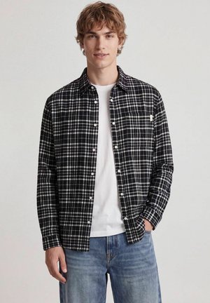 Ung mand iført en sort og hvid ternet skjorte over en hvid t-shirt og blå jeans, stående med den ene hånd i lommen.