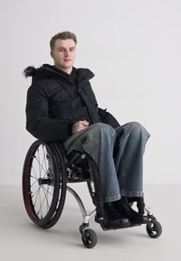 Chaqueta acolchada negra con capucha forrada de piel, llevada sobre una camiseta negra. Jeans azules holgados y botas negras. Usuario sentado en una silla de ruedas manual.