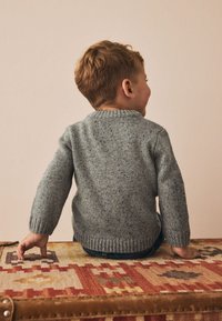 Grå stickad tröja med ett strukturerat mönster, ribbade mudd och halslinning, bärs av ett barn som sitter på en mönstrad textilyta.