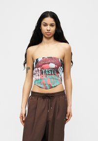 Jeune femme aux longs cheveux foncés portant un haut graphique sans bretelles avec des lèvres et du texte, associé à un pantalon brun ample à cordon de serrage.