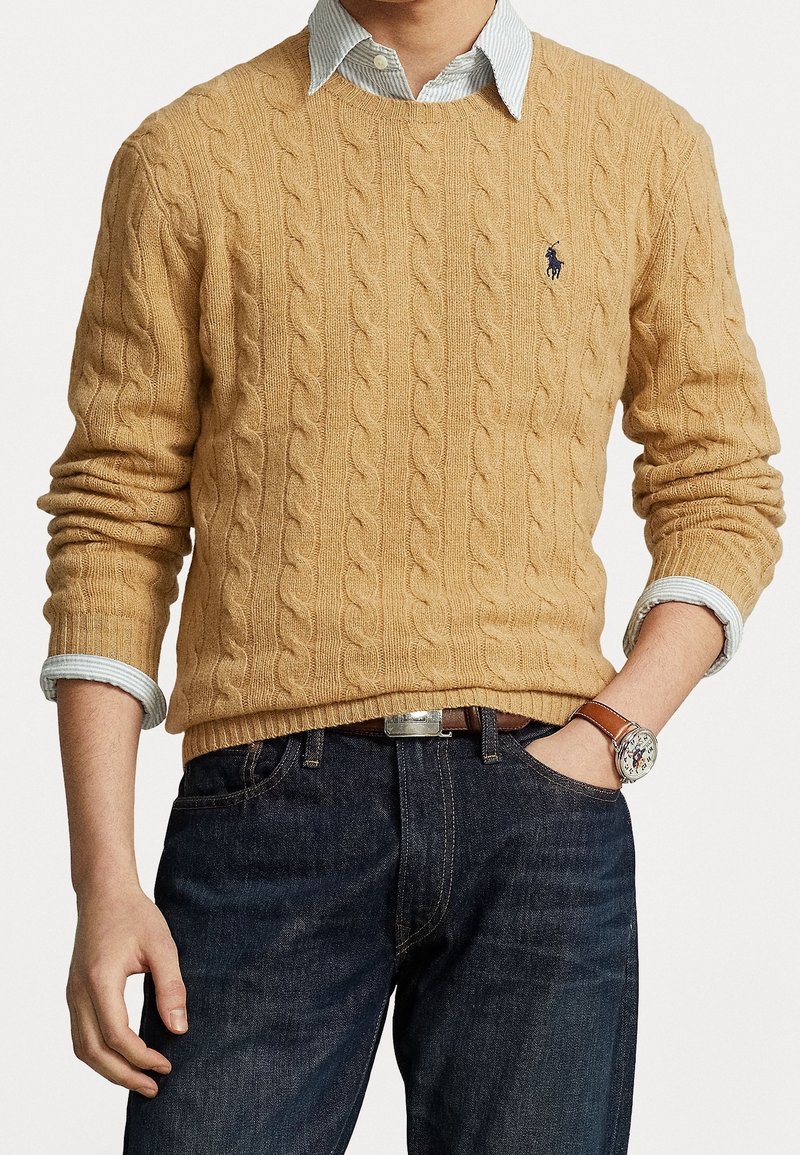 Homme portant un pull en maille torsadée beige sur une chemise rayée bleu clair, avec un jean foncé et une montre en cuir marron, une main dans la poche.