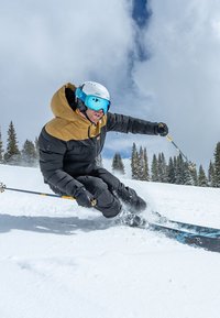 Skifahrender Mann in einer schwarz-braunen isolierten Jacke, mit einem weißen Helm und blauen Skibrillen, der durch den Schnee mit Stöcken carve. Kiefernbäume im Hintergrund.