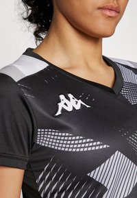 T-shirt de sport noir avec des motifs géométriques gris, un design en V et un logo blanc sur l'épaule gauche. Texture lisse, forme ajustée.