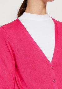 Rosa cardigan med V-halsdesign, tillverkad av slätt tyg, med en framknäppning och subtila sömnadsdetaljer. Buren över en vit topp.