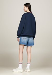 Felpa oversize blu navy con spalle basse, abbinata a pantaloncini di jeans blu. Sneakers bianche e calzini a righe completano l'outfit.