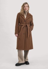 Brauner Mantel aus Wollmischung mit breiten Revers, tailliert mit Gürtel und kontrastierenden Knöpfen; kombiniert mit beigen Cordhose und schwarzen Plateauschuhen.