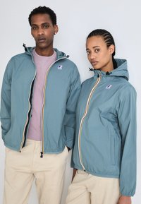 Dos personas llevan chaquetas ligeras a juego en color azul verdoso con capuchas, con cremalleras en contraste y un logotipo en el pecho. Pantalones neutros complementan el conjunto.