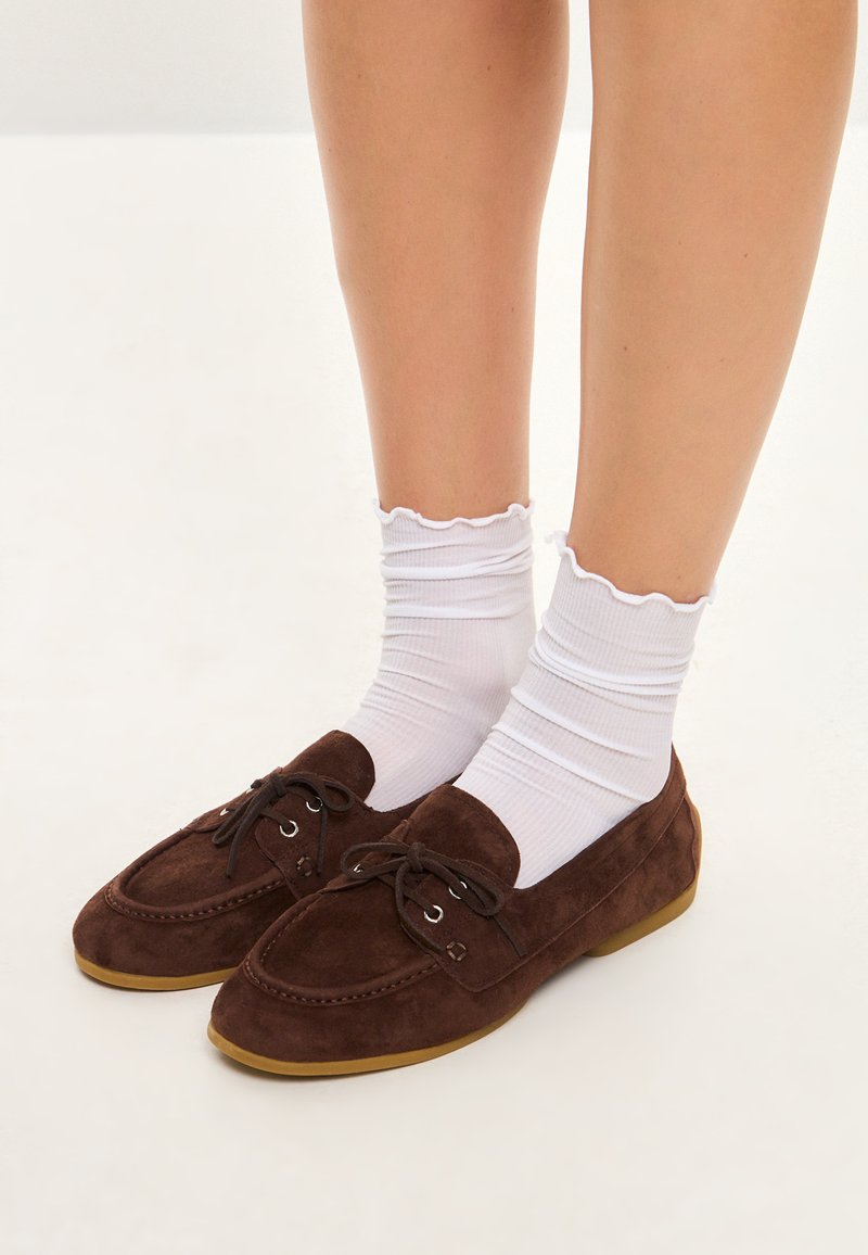 Brune ruskind loafers med rund tå og snørebåndsdetaile, parret med hvide ribbede sokker med en flæsekant. Flad gummisål.