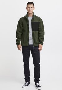 Grønn fleecejakke med svart brystlomme, glidelås, brukt over en lys grå skjorte. Svarte jeans og svarte sneakers fullfører antrekket.