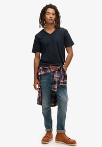 Donkerblauw v-hals t-shirt, lichtblauwe jeans, bruine veterschoenen en een rood geruit overhemd om de taille geknoopt. Casual pasvorm en gelaagde uitstraling.