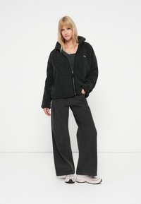 Schwarze Fleecejacke mit Reißverschluss und hohem Kragen, kombiniert mit grauen weit geschnittenen Hosen und hellen Sneakern. Strukturiertes, gemütliches Material.