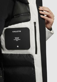 Zwart-wit pufferjackinterieur met een groot zwart label met de tekst "KHUJOTM" en een ritszak aan de linkerkant.