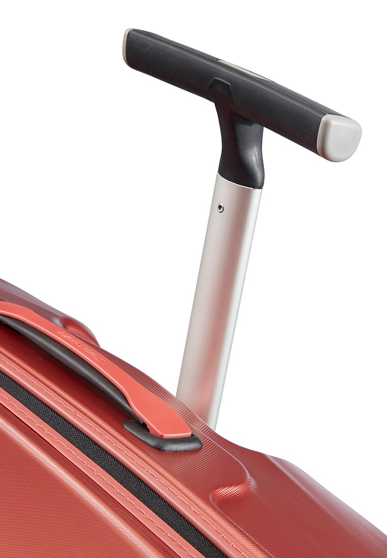 Maleta rígida roja con un mango telescópico plateado, superficie texturizada y un acento moldeado. Presenta un asa superior para un fácil manejo.