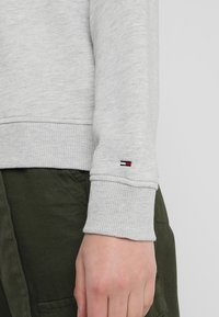 Jasnoszara bluza z prążkowanymi mankietami; małe haftowane logo w czerwonym, białym i niebieskim kolorze na rękawie; połączona z zielonymi spodniami z tkaniny.