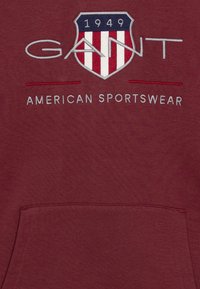 Felpa con cappuccio color marrone scuro con logo ricamato bianco e blu navy, design a scudo, con scritte "GANT 1949" e "AMERICAN SPORTSWEAR" sopra una tasca frontale.