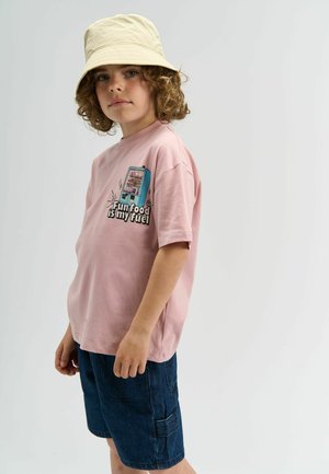 Roza pamučna majica s grafikom automata za prodaju i natpisom "Zabavna hrana je moje gorivo", uparen s tamnoplavim hlačama do koljena i bež šeširom.