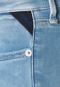 Jeans de denim azul claro con una etiqueta de tela negra con "REPLAY" bordado en azul. Detalle de botón de metal plateado en la cintura.