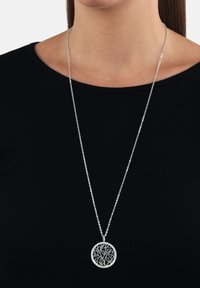 Silver tree of life-hänge på en tunn kedja. Hängsmycket har en intrikat utskuren design och är cirkulärt. Bärs över en svart skjorta.