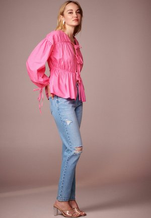 Blouse rosa con maniche a sbuffo e dettagli legati, abbinata a jeans chiari strappati e sandali con tacco trasparente. Contrasto di colore morbido.
