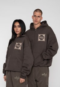 Brune zip-up hættetrøjer har forlommer og et dekorativt broderet design på brystet. Begge bærer matchende cargo bukser i en mørkere brun.