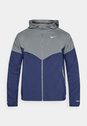 Nike dzseki kapucnival, szürke texturált felső résszel és tengerészkék alsó résszel. Reflektív elemekkel és teljes cipzárral rendelkezik.