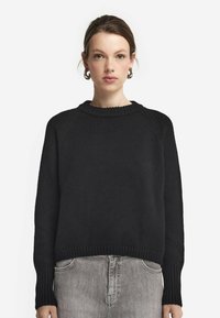 Svart stickad tröja med rund hals, ribbade ärmslut och nederkant. Har en avslappnad passform och strukturerad tyg, matchad med grå denimbyxor.