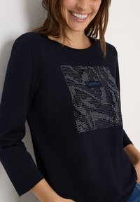 T-shirt à manches longues marine en tissu doux, arborant un motif géométrique de clous métalliques et un logo bleu au centre.