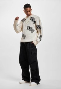 Creme sweater med sorte "DEF" logoer, afslappet pasform og ribbede manchetter, parret med løse sorte cargo bukser og hvide sneakers.