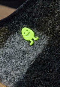 Tissu feutré avec un dégradé de gris et de noir, présentant un petit personnage souriant en plastique vert attaché à la surface.