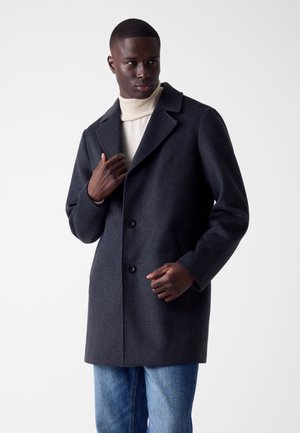 Manteau en laine gris foncé avec un col cranté, deux boutons et des poches latérales, porté sur un col roulé crème et un jean bleu.