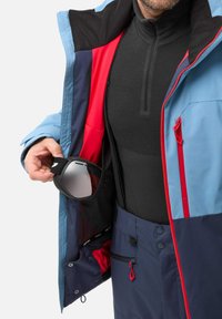 Ski jacket with a blue outer layer, red accents, and a black inner lining. It has zippered pockets and a hand holding goggles.