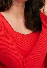 Donna che indossa un maglione rigato rosso brillante con maniche lunghe, capelli castano scuro visibili e collo scoperto.