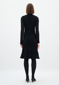 Rochie neagră din dantelă, cu mâneci lungi și guler înalt, având un tiv evazat și un model texturat. Purtați cu colanți negri și pantofi.